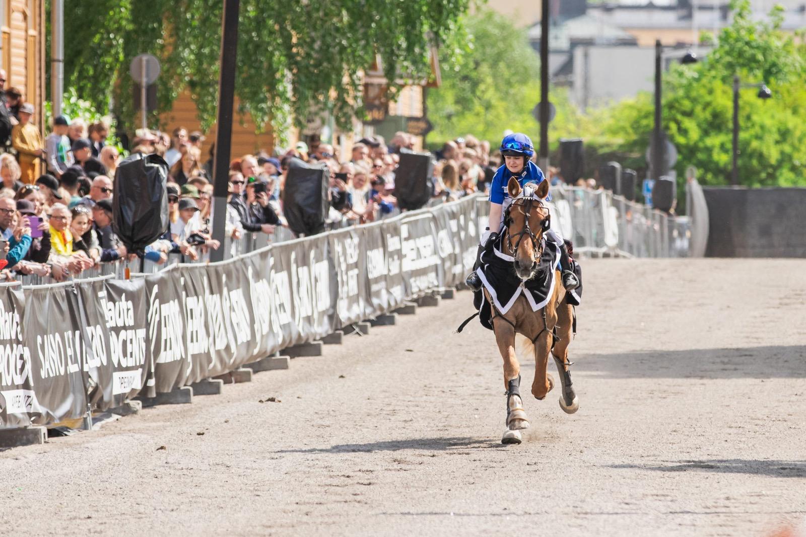 Ihminen ratsastaa hevosella, sivussa aidan takana ihmisjoukko katsomassa.